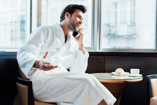 happy bearded man in white bathrobe talking on smartphone near coffee table with breakfast - Powered by Adobe