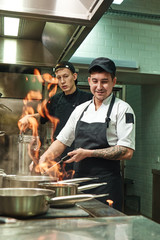 Working with fire. Two young and positive cooks learning how to make a flambe on food in restaurant kitchen
