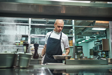 I should be careful. Serious male chef with several tattoos on his arms holding a frying pan above the oven in a restaurant kitchen