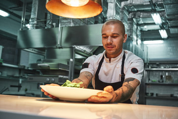 Ready to eat. Handsome smiling chef with tattoos on his arms, in black apron holding ready dish in modern restaurant kitchen.