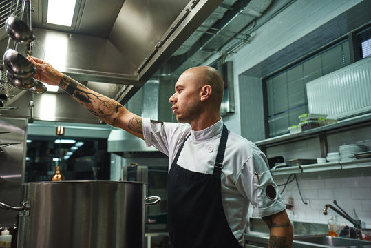Famous soup. SIde view of attractive bald chef in apron taking a ladle for soup cooking while standing in a restaurant kitchen