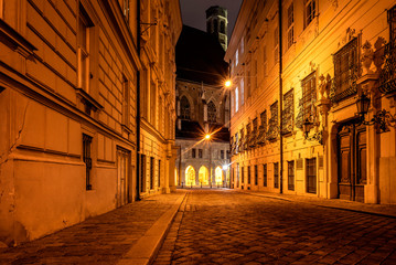 Fototapeta na wymiar Austria, Vienna, Metastasiogasse: Narrow alleyway street with cobblestone, old houses, lights and famous Minoriten church in the background at dark night in the city center of the Austrian capital.