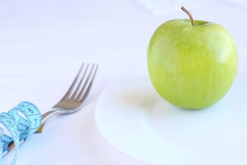 Fresh organic green juicy apple on white plate with blurred fork and knife and blue measuring tape on wooden neutral background. Diet concept with raw vegetarian food. Green fruit at the dish