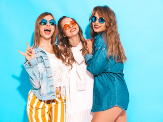 Three young beautiful smiling hipster girls in trendy summer casual jeans clothes. Sexy carefree women posing near blue wall. Positive models going crazy in sunglasses
