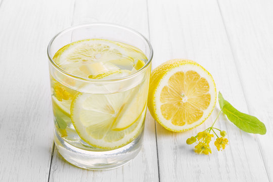 Refreshing Drink For Health, Lemon Water In A Glass And Yellow Linden Flowers On A White Table.