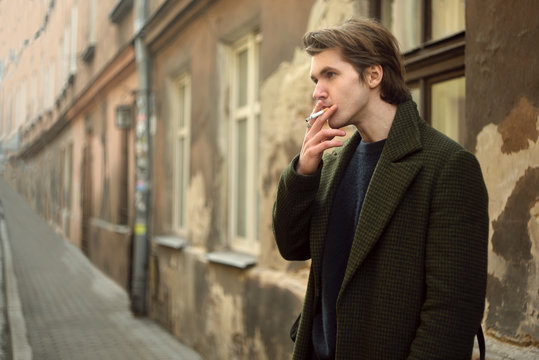 Man on the old street smoking cigarettes, in a long coat. Man befriends a cigarette in the city.  A man like from old movies, vintage.