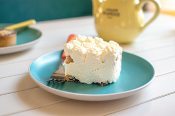 Сream cake on blue plate with fork, yellow tea pot and green cup. Delicious white dessert and tea in cafe. A piece of coconut cake
