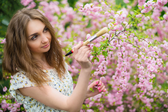 .A beautiful fair-haired girl stands in the garden of a blooming pink Sakura with her eyes closed and paints a brush of pink eyes. Horizontal photography.