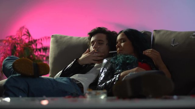Drunk couple smoking cigarettes on nightclub couch, hangover girl feeling sick