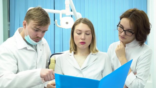 Group of interns or young doctors in white lab coats looking at blue folder, medicine and health care concept. Three dentists in the cabinet discussing disappointing diagnosis, lookig sad.