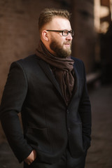 Obraz premium A man with a beard in a black warm coat with a scarf poses to advertise men's clothing in the winter. Advertise men's clothing