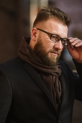 Fototapeta premium A man with a beard in a black warm coat with a scarf poses to advertise men's clothing in the winter. Advertise men's clothing