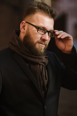 Fototapeta premium A man with a beard in a black warm coat with a scarf poses to advertise men's clothing in the winter. Advertise men's clothing