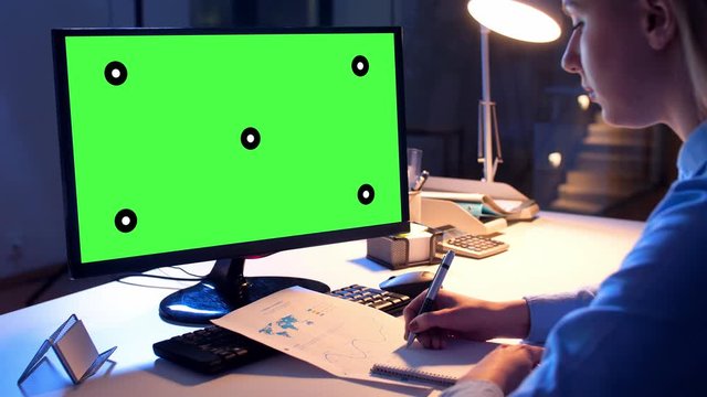 technology, virtual effects and business concept - businesswoman with chroma key green screen on computer monitor writing to notebook at night office