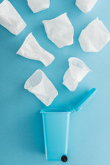 top view of white plastic cups falling down toy trash can on blue background