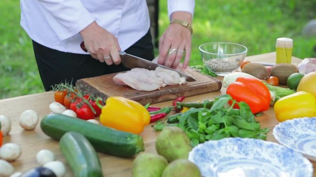 Healthy summer food. Cook cuts the chicken on a wooden board. Table with different vegetables. Mushrooms and red tomatoes. Green pepper and salad. Happy people having camping and bbq party outdoor.