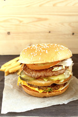 Big classic american burger with two meat cutlets tomato, onion, lettuce, cheese on kraft paper with french fries on wood.  Unhealthy food