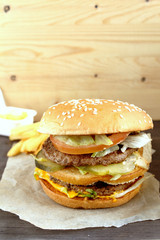 Big classic american burger with two meat cutlets tomato, onion, lettuce, cheese on kraft paper with french fries on wood.  Unhealthy food