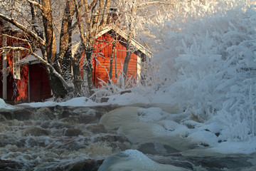 Frozen rapid flowing.  Sun shines over old red house in a sunny January day