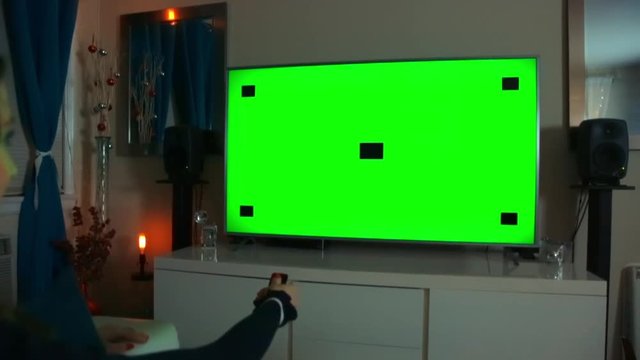Slow Motion Dolly Shot of a caucasian woman sitting comfortably in her living room, watching TV at night, and uses a remote to press a button. The TV screen is set to green screen with tracking marks.