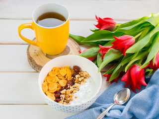 Delicious yogurt bowl with corn flakes, nuts and jam on a white wooden table. Healthy and organic nutrition concept. Tulips with cup of tea and breakfast.