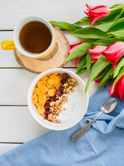 Delicious yogurt bowl with corn flakes, nuts and jam on a white wooden table. Healthy and organic nutrition concept. Tulips with cup of tea and breakfast. Top view