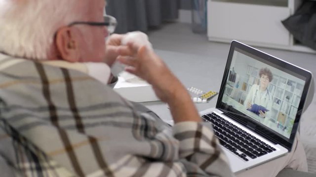 Rare View Of Sick Old Man Sitting On Couch In Living Room With Laptop On His Knees And Telling Female Doctor About His Sickness