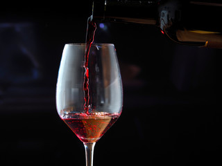 Red wine is poured into a large wine glass from a dark bottle on a black background.