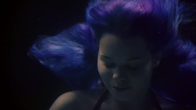young woman with blue hair is keeping afloat underwater in darkness, opening eyes