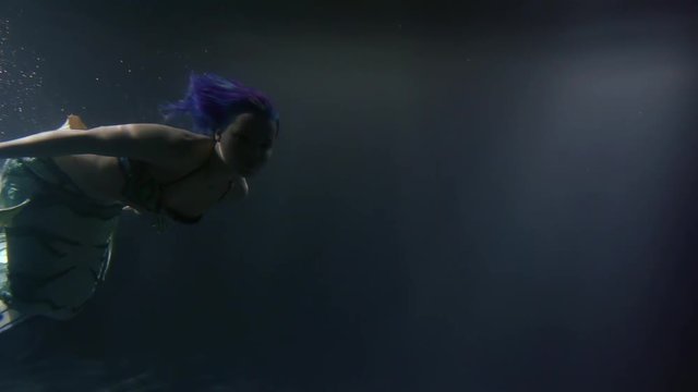 young graceful mermaid with blue hair is swimming in ocean water in night time, swaying tail