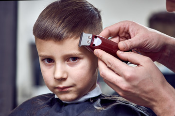 Barber making a haircut to a cute European boy using cutting machine.