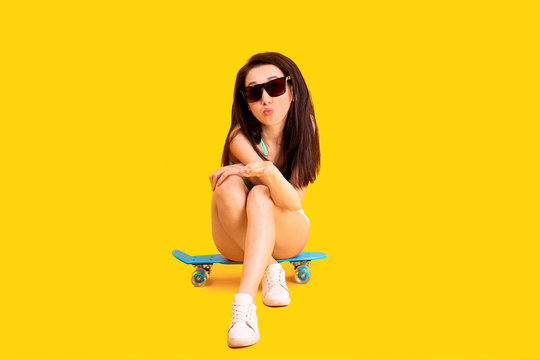 beautiful girl in sunglasses, sitting on a skateboard and sending air kiss, image on yellow background