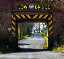 The reflective yellow and black warning signs around a railway bridge highlighted over an empty road, with a view up the tree lined hill to a vehicle in the distance. - 256913356