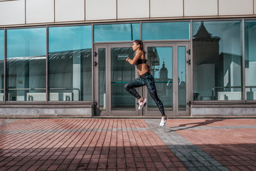 Obraz na płótnie Canvas Girl morning jog summer city, listening music headphones. Runs high jump. Active lifestyle, lifestyle. Free text space for motivation. On background of glass windows. Clothing leggings top, sneakers.