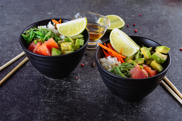 Two homemade healthy Poké bowl with rice, salmon, avocado, white onion, scallion greens, sesame oil, soy and sesame sauce, seaweed (wakame), lime and rice vinegard on a dark backgound, closeup