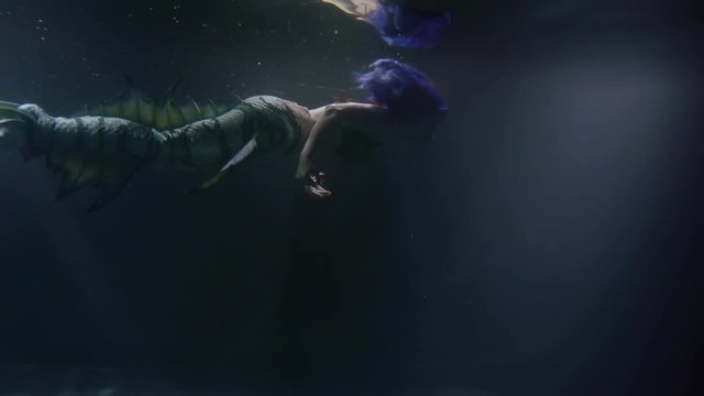 amazing mermaid is floating underwater in darkness, approaching to surface