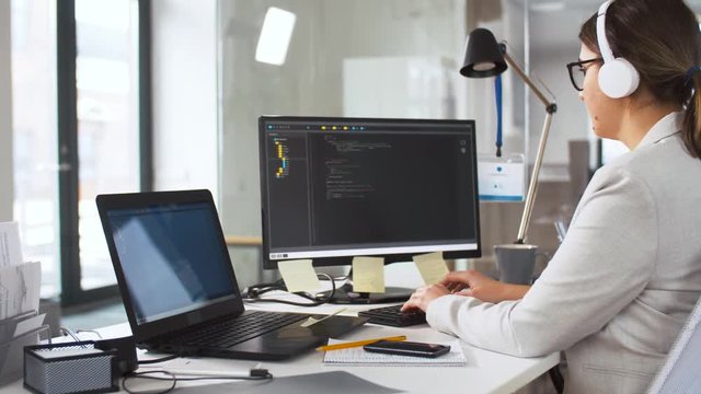 business, programming and technology concept - female programmer in headphones working with script on computer screen and listening to music at office