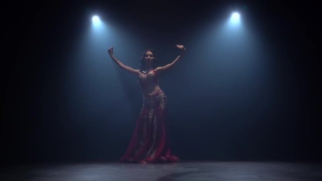 Girl starts dancing belly dance in dark studio. Black smoke background. Slow motion