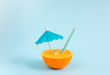 Orange juice with tubule and umbrella. Orange cut in half on pastel blue background. Minimal summer concept. Flat lay