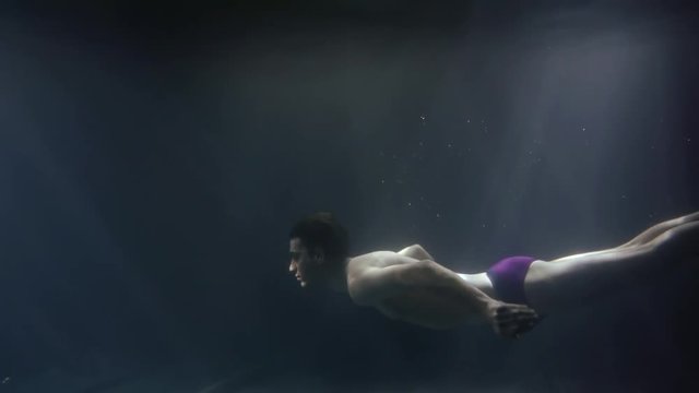 young slim man dressed swimming trunks and red socks is swimming underwater in darkness