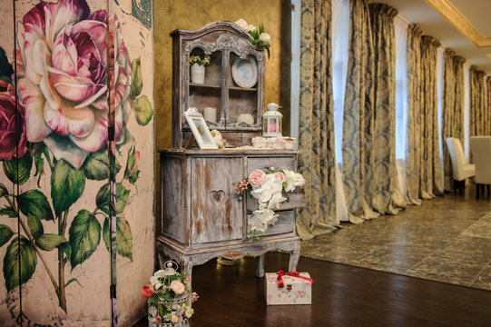 close up photo of a wedding decor: a vintage wooden cupboard decorated with flower. photo frame, candles, lamps, jewerly box near a retro parvan with roses motives