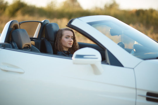 beautiful sexy girl with long hair in a leather jacket and leather pants in sunglasses sits in an expensive car at sunset