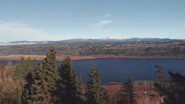 Aerial drone shot of the Columbia River Gorge on a beautiful morning sweeping past evergreen trees into a vast wide angle view of the river with snow covered peaks in the distance