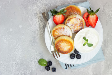 Cottage cheese fritters (syrniki) served with sour cream, fresh berries (strawberry and blueberry) and mint on a light grey background. Healthy breakfast or diet lunch. Copy space.