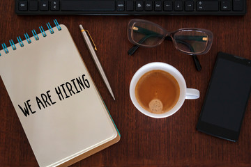 Office desk with computer, coffee cup, smartphone, glasses and We are hiring written on notebook