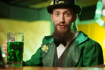 Dark-haired bearded young man in a green costume with a shamrock symbol sitting at the table
