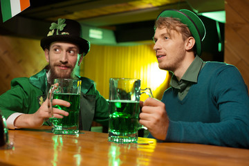 Dark-haired bearded young man in a leprechaun hat and his friend looking interested