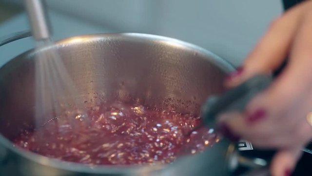 close view woman with purple manicure stirs red sweet boiling caramel in metal cooking saucepan using whisk