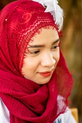 Women muslim, women islamic,wearing a red fabric head covering adorn with white fabric flowers wear shirt white sit poses photography on the chair under the tree before to ramadan month.