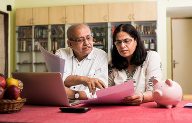 Senior Indian/asian couple accounting, doing home finance and checking bills with laptop, calculator and money while sitting on sofa/couch at home